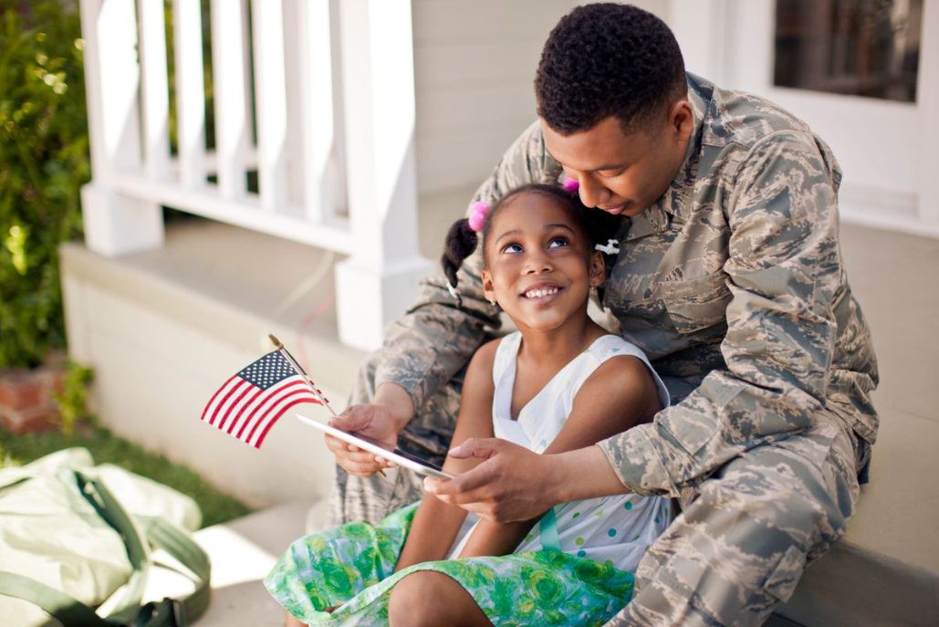 A father in his military uniform sitting on the front stairs of a house with his daughter.